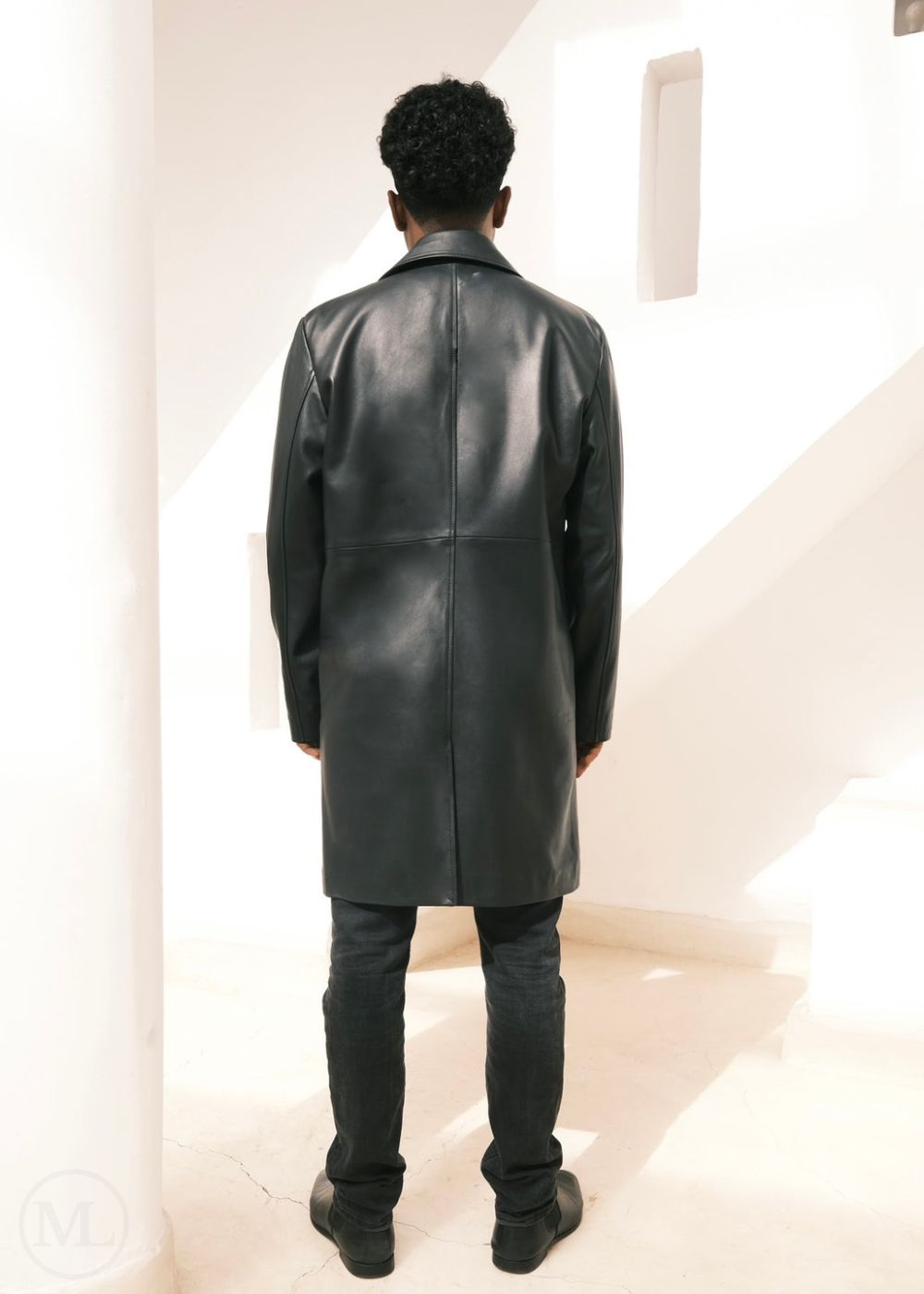 Man wearing a black leather trench coat standing in a minimalistic room.