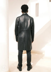 Man wearing a black leather trench coat standing in a minimalistic room.