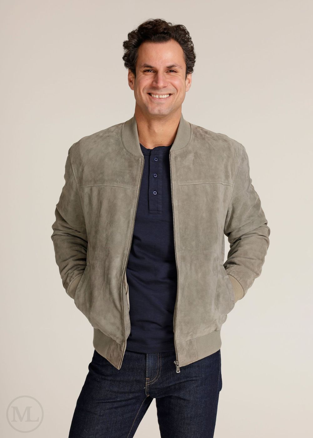 Man wearing a light gray suede jacket over a navy shirt and dark jeans on a beige background