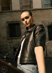 Women wearing a cropped black leather jacket in shirt style and jeans standing in front of a stone building.