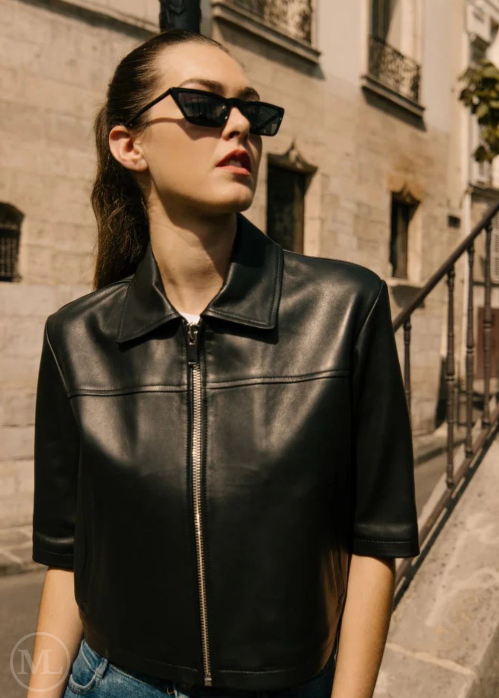 Woman wearing a cropped black leather shirt style jacket and sunglasses on a city street.