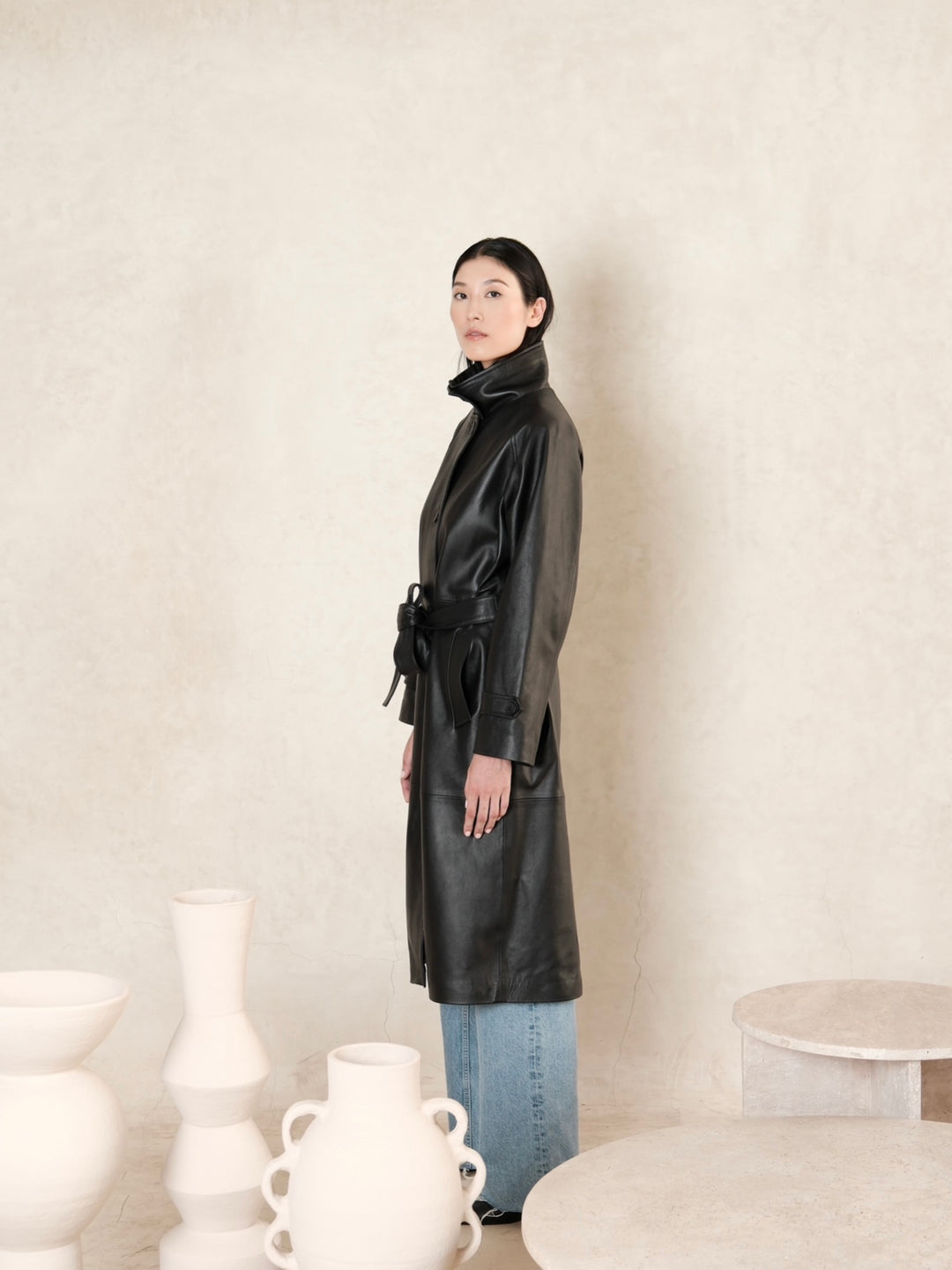 women wearing a black leather trench coat, standing in side postiion, to show left side of the coat