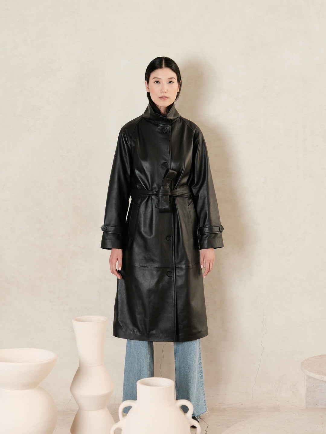 women wearing a black leather trench coat with belted waist and a beige background