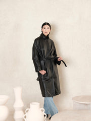 women wearing a black leather trench coat holding a black belt against a beige wall