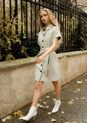 Woman in a stone beige real leather dress standing on a sidewalk with a black metal fence and greenery in the background.