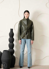 Women wearing a green leather coat style jacket and blue jeans standing against a white wall with decorative vases.