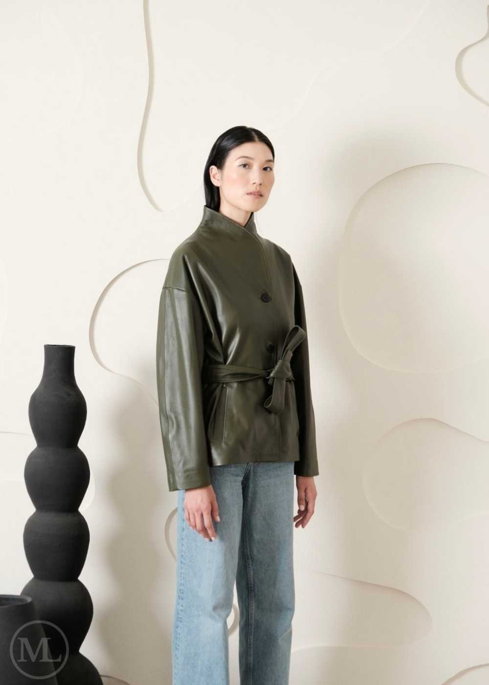 Woman wearing a green leather coat, and light blue jeans standing against a white wall with decorative elements.