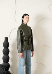 Woman wearing a green leather coat, and light blue jeans standing against a white wall with decorative elements.