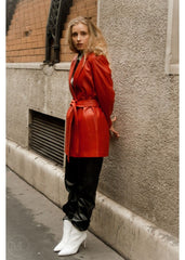 Woman in a red leather blazer coat and black pants standing against a textured wall.