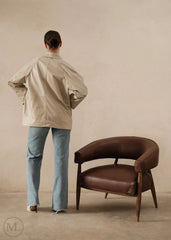 Women  wearing a lvory leather blazer  jacket and light blue jeans standing next to a brown leather chair against a beige wall.