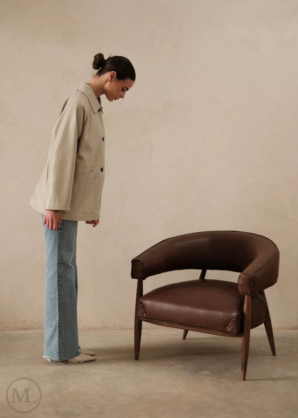 Woman in a lvory leather blazer coat and jeans standing next to a brown leather chair against a beige 