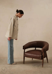 Woman in a lvory leather blazer coat and jeans standing next to a brown leather chair against a beige 