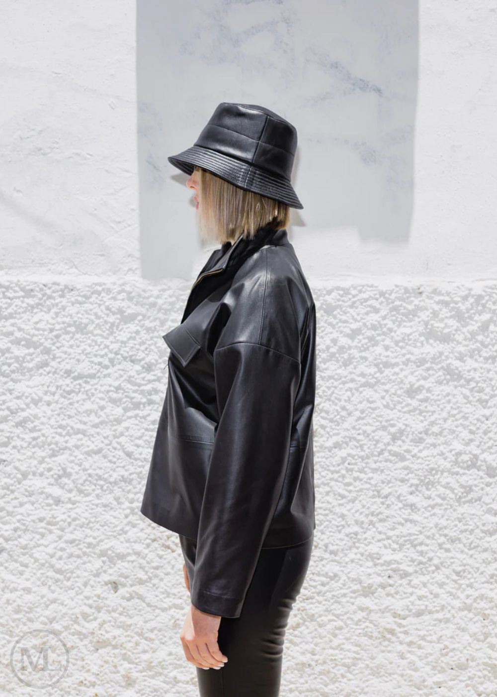 Women wearing a black leather jacket and hat against a textured white wall.