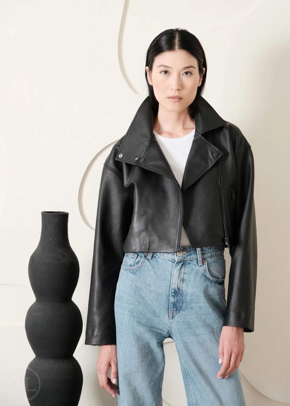 Women wearing a Cropped black leather Biker jacket and blue jeans standing next to a black vase on a white background