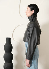 Women wearing a Cropped black leather Biker jacket and light blue jeans standing next to a black vase on a white background