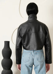 Women wearing a Cropped black leather Biker jacket and blue jeans standing next to a black vase on a white background
