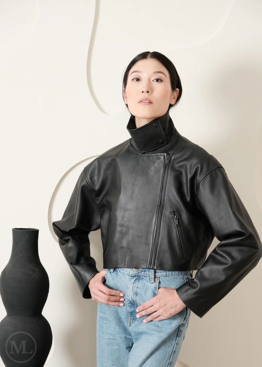 Women wearing a Cropped black leather Biker jacket and light blue jeans standing next to a black vase on a white background
