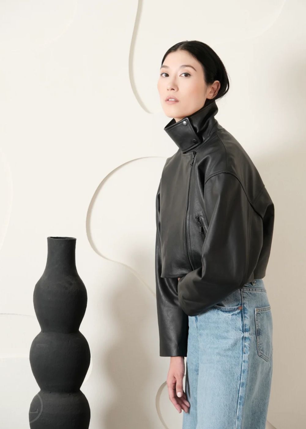 Women wearing a Cropped black leather Biker jacket and light blue jeans standing next to a black vase on a white background
