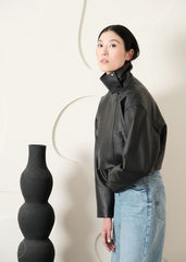 Women wearing a Cropped black leather Biker jacket and light blue jeans standing next to a black vase on a white background