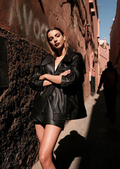 Woman in a black leather biker jacket standing against a textured wall in an alley.