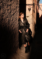 Woman in a black leather trench coat standing in a narrow alleyway.