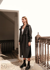 Woman in a long black leather trench coat standing on a balcony with architectural details.