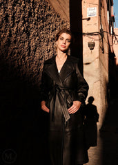 Woman in a black leather trench coat standing in a narrow alleyway with warm lighting.