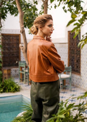 Woman wearing a brown leather jacket and green pants standing by a pool with plants around.