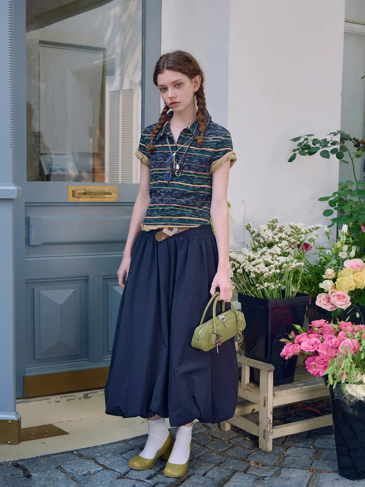 Woman in a striped shirt and navy skirt standing in front of a blue door with flowers in the background.