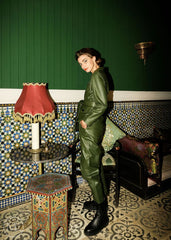 Woman wearing a leather coat standing in a room with decorative tiles and a lamp.