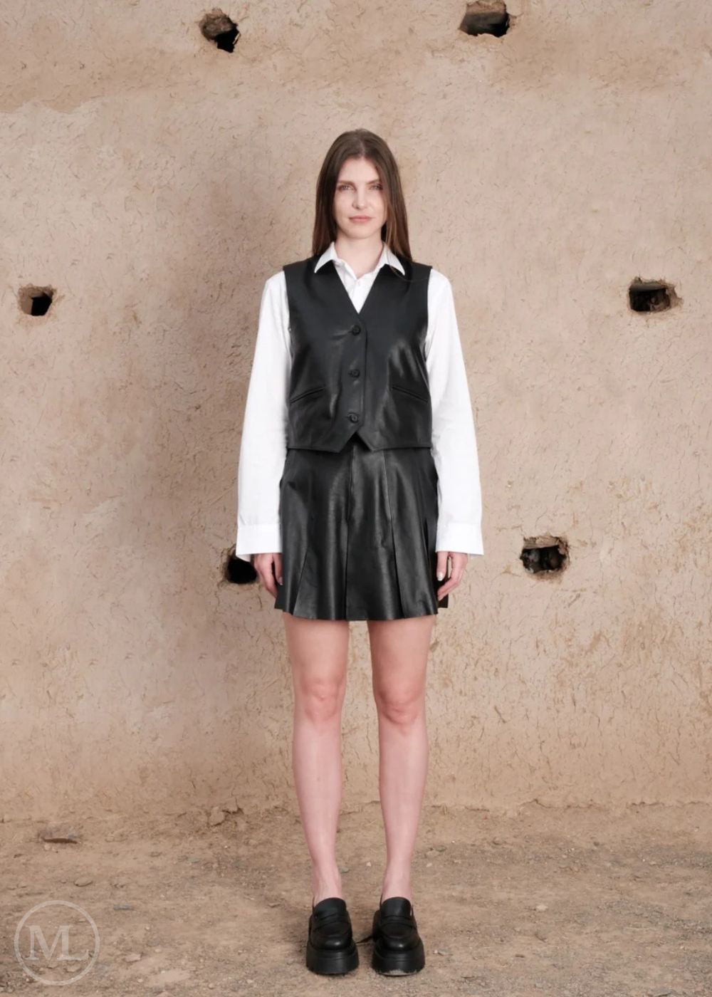 Women wearing a black leather vest and skirt against a textured beige wall.