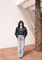 Women wearing a Cropped black leather Biker jacket and light blue jeans standing against a neutral wall with palm trees.