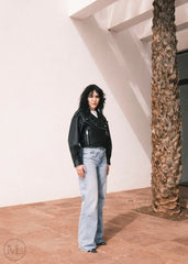 Women wearing a Cropped black leather Biker jacket and light blue jeans standing against a neutral wall with palm trees.