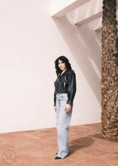 Women wearing a Cropped black leather Biker jacket and light blue jeans standing against a neutral wall with palm trees.