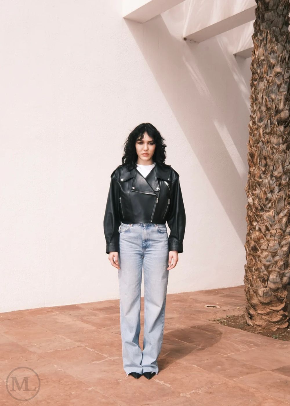 Women wearing a Cropped black leather Biker jacket and light blue jeans standing against a neutral wall with palm trees.