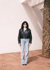 Women wearing a Cropped black leather Biker jacket and light blue jeans standing against a neutral wall with palm trees.