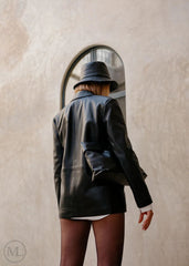 Woman wearing a black blazer Coat, white shirt, and hat standing against a beige wall.