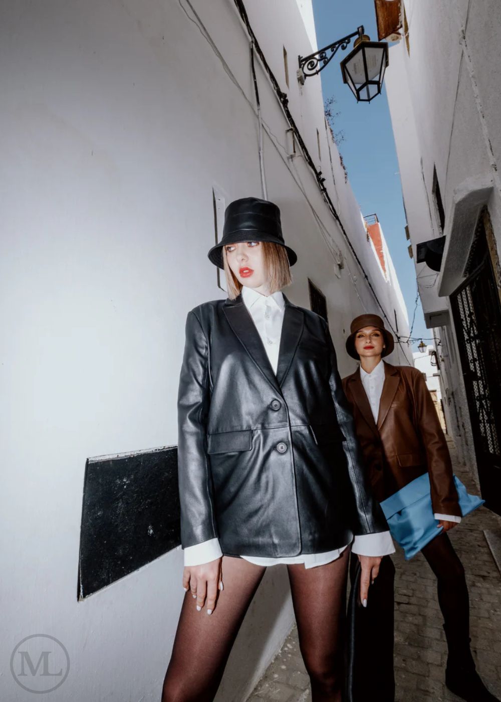 Woman wearing a black blazer Coat, white shirt, and hat standing against a beige wall.