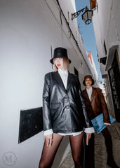 Woman wearing a black blazer Coat, white shirt, and hat standing against a beige wall.