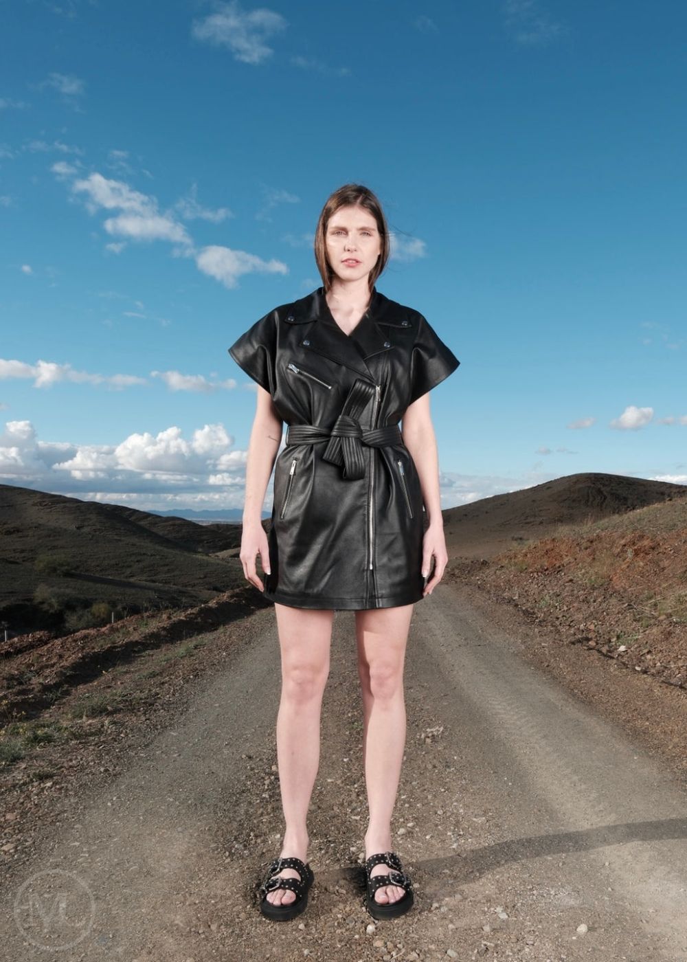 Woman in a black leather dress  in biker style standing on a desert road with a blue sky.